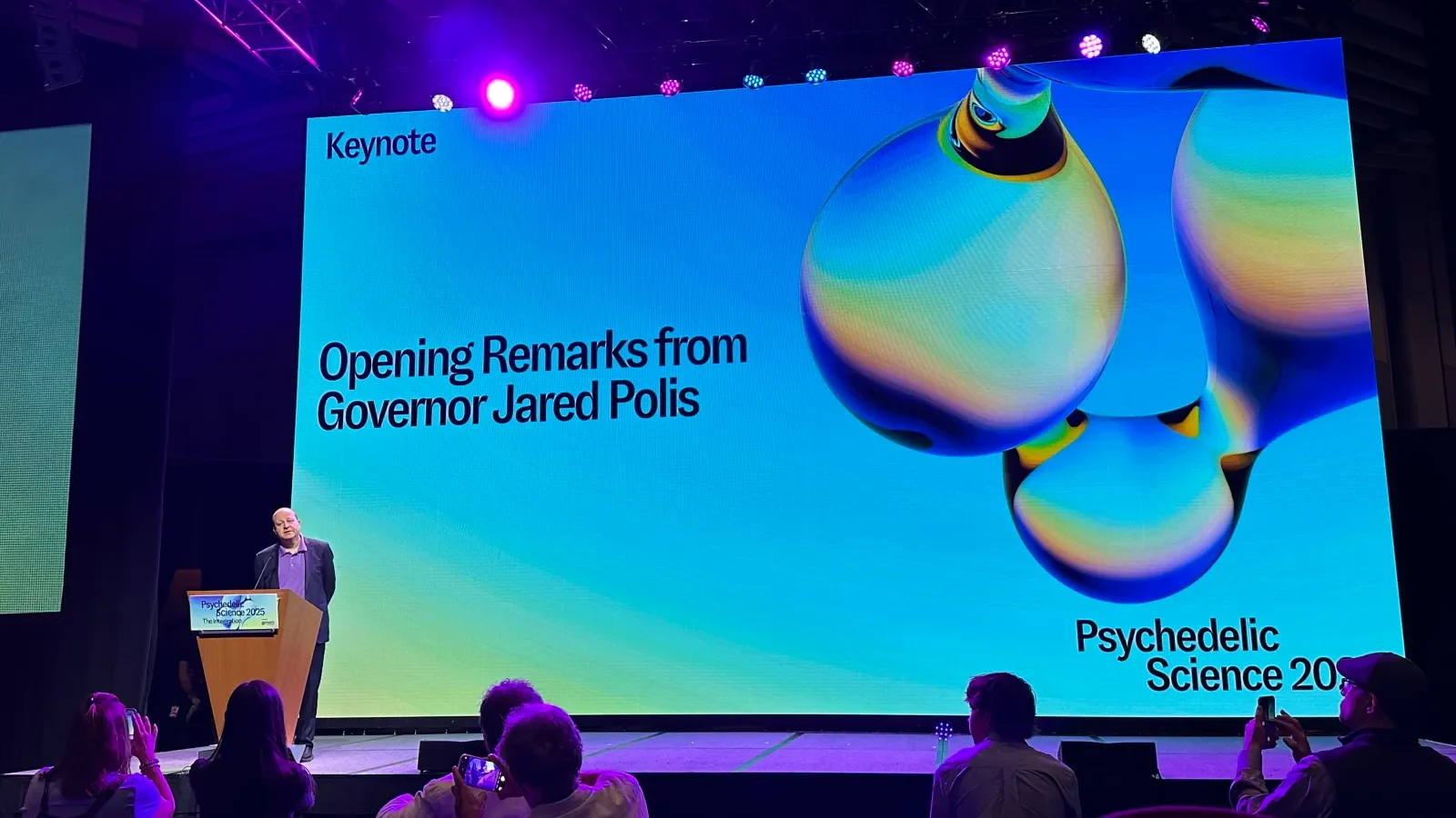 The keynote stage: Opening Remarks from Governor Jared Polis on the screen beside three iridescent molecules, the Psychedelic Science 2025 lockup in the corner, the governor at the podium
