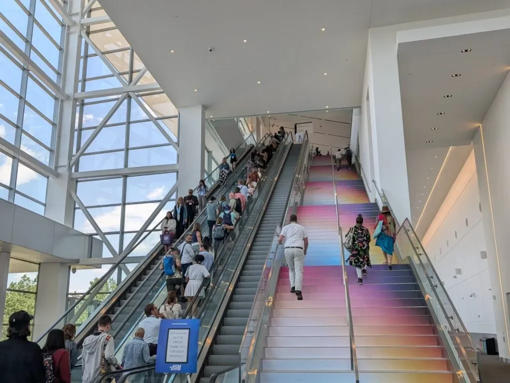 Attendees riding the escalator beside the grand stair wrapped in the pink-to-blue gradient